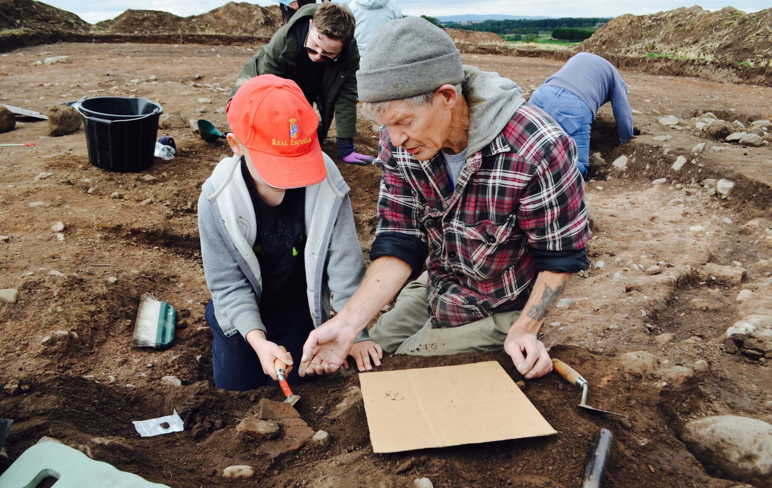 A child wearing a red hat is being shown an archaeological artefact by an older adult wearing a grey beanie on an archaeological dig site