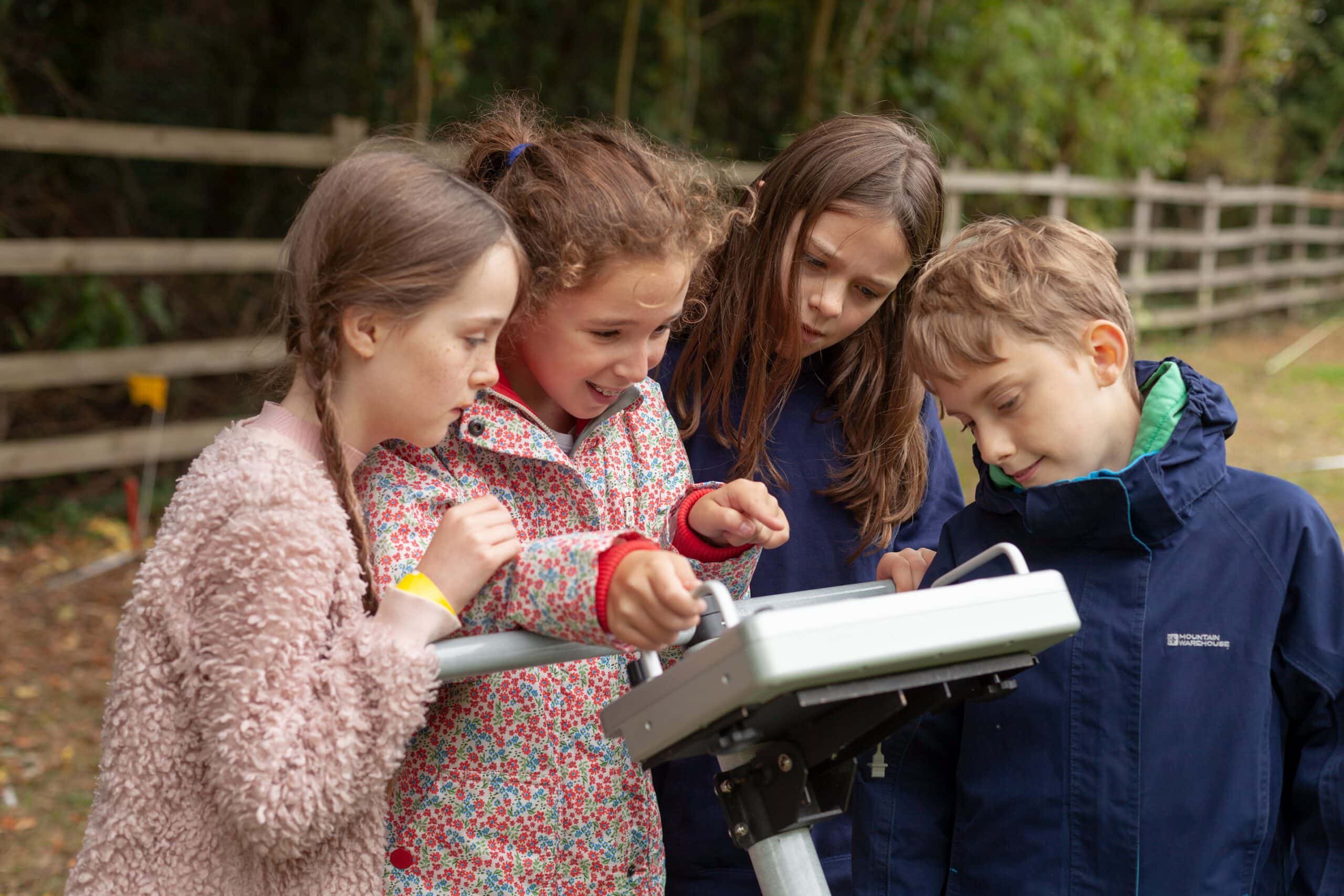 A group of children looking at a screen attached to geophysical equipment in a filed with a fence.