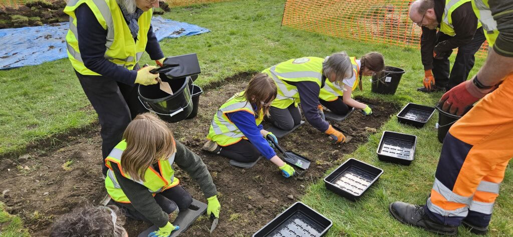 A group of young archaeologists wearing high-vis vests digging in an archaeological trench.