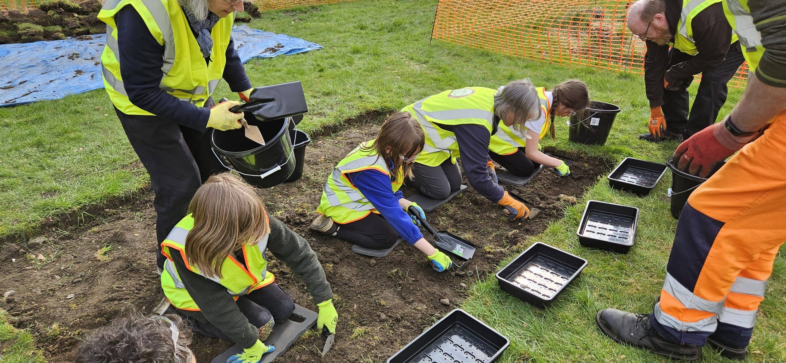 A group of young archaeologists wearing high-vis vests digging in an archaeological trench.