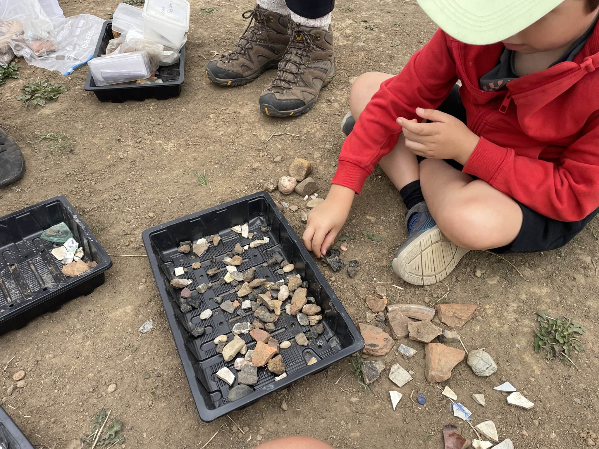 A child sat on the ground sorting through small pieces of bone, pottery and stone in a black tray.