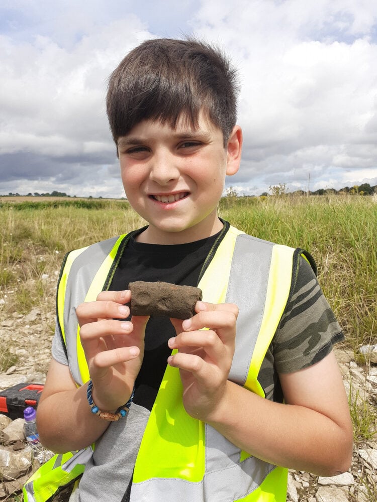 A boy wearing a high-vis vest stands in a field on an archaeological dig holding a small brown piece of pottery with finger prints on it.