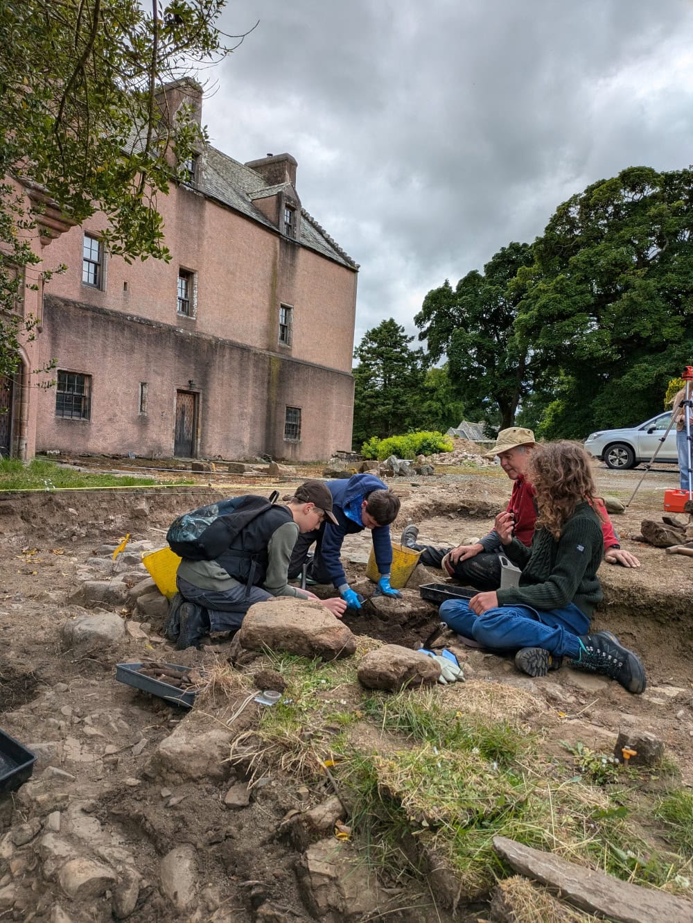 Three young people and one adult sat outside around a shallow trench on an archaeological dig outside a large rural house.