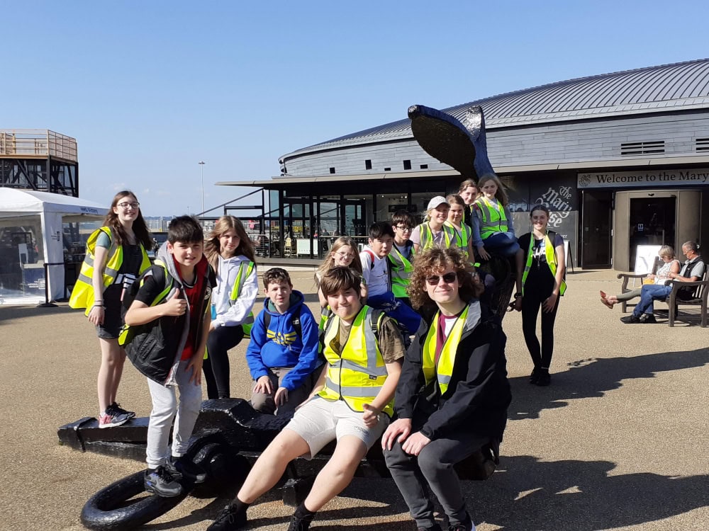 A group of 17 children and adults stand in front of the entrance to a museum. The museum sign reads 'The Mary Rose'.