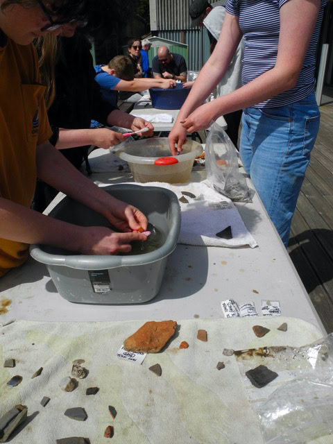 A group of young people gather around a long table on which are washing up bowls of water. They use toothbrushes to wash archaeological objects such as tiles and bricks.