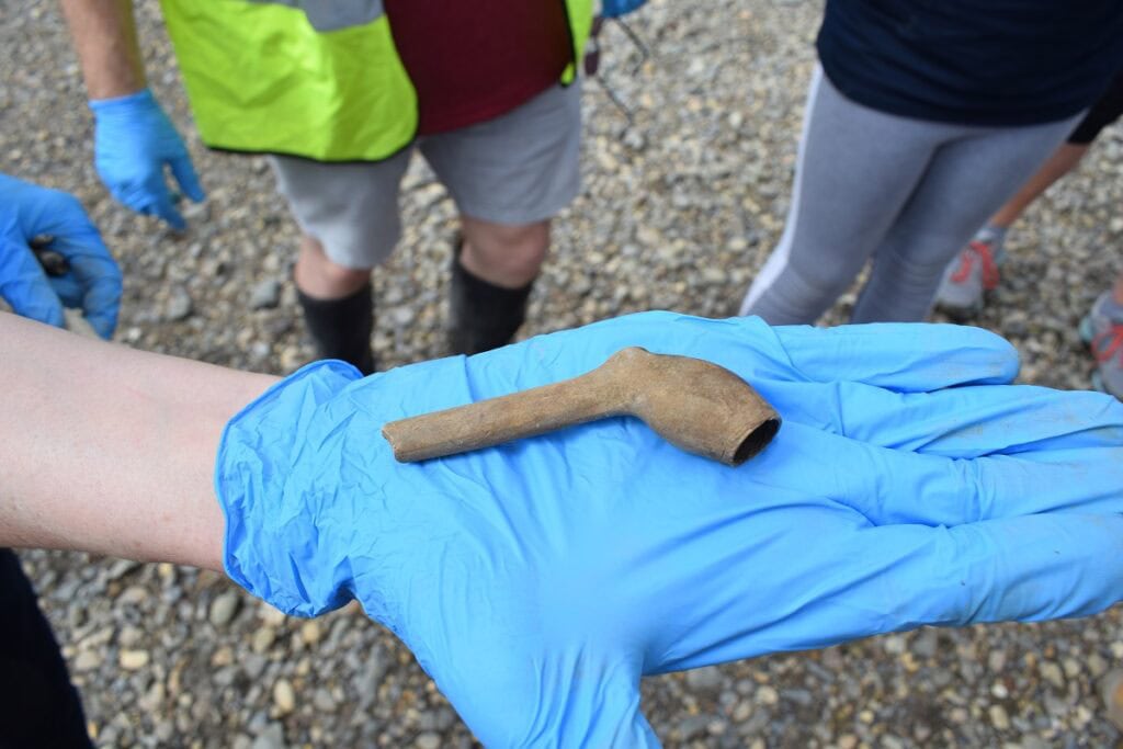 A clay smoking pipe is held in a gloved hand on the banks of a river.