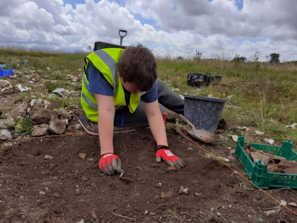 A boy leans over the edge of a dig pit in a field with a trowel.