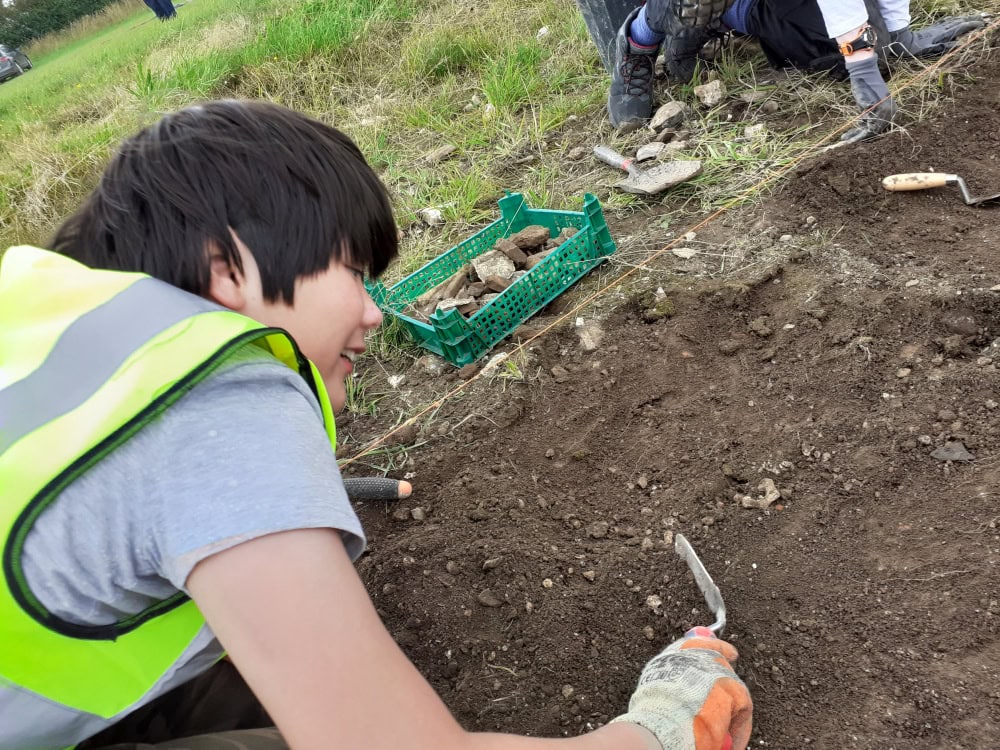 A young person kneels on the ground to excavate an archaeological dig with a trowel.