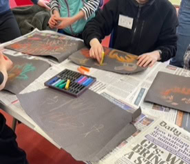 Young people sat at a table drawing handprints onto black paper using colourful crayons to imitate cave art.