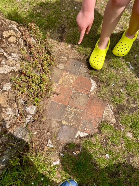 Some old red and grey tiles, some with gold-coloured designs, are visible on the ground through some grass. A young person's feet and legs can be seen next to the tiles and their hand points to them.