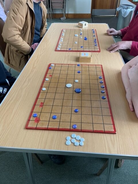 Young people sat around a table play the Roman boardgame 'Aquila' on wooden boards with blue and white counters.