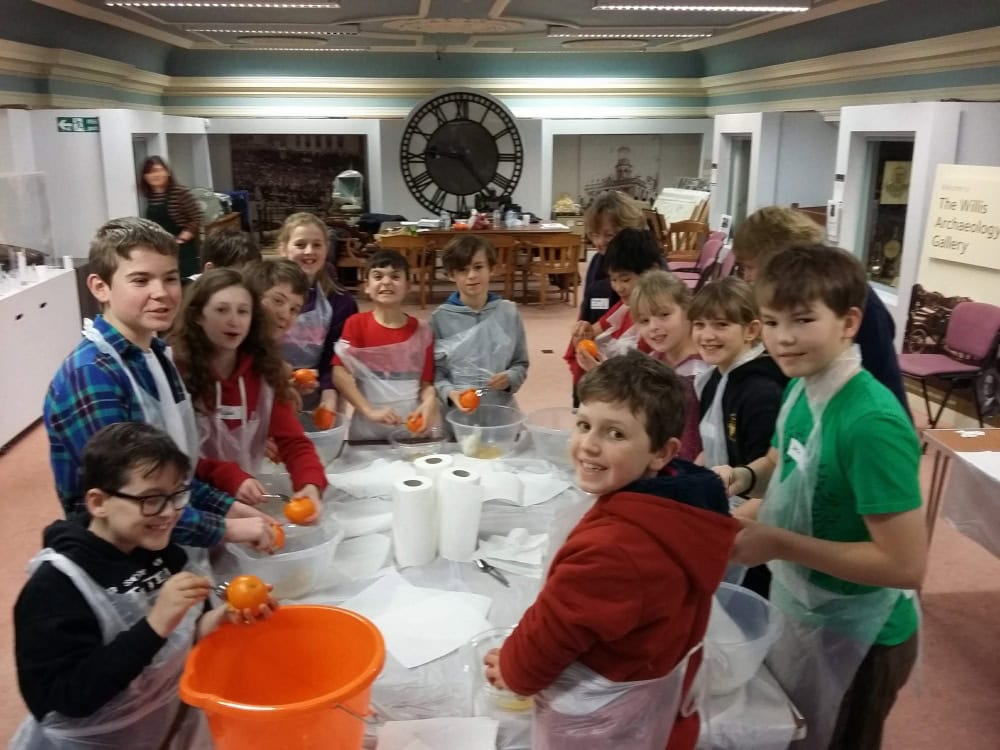 A group of 13 children wearing plastic aprons and holding oranges stand around a table covered in paper towels.