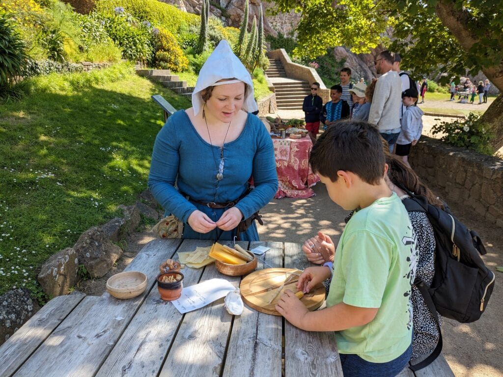 Two children in modern clothes talking to a woman in medieval clothes and holding some beeswax candles.