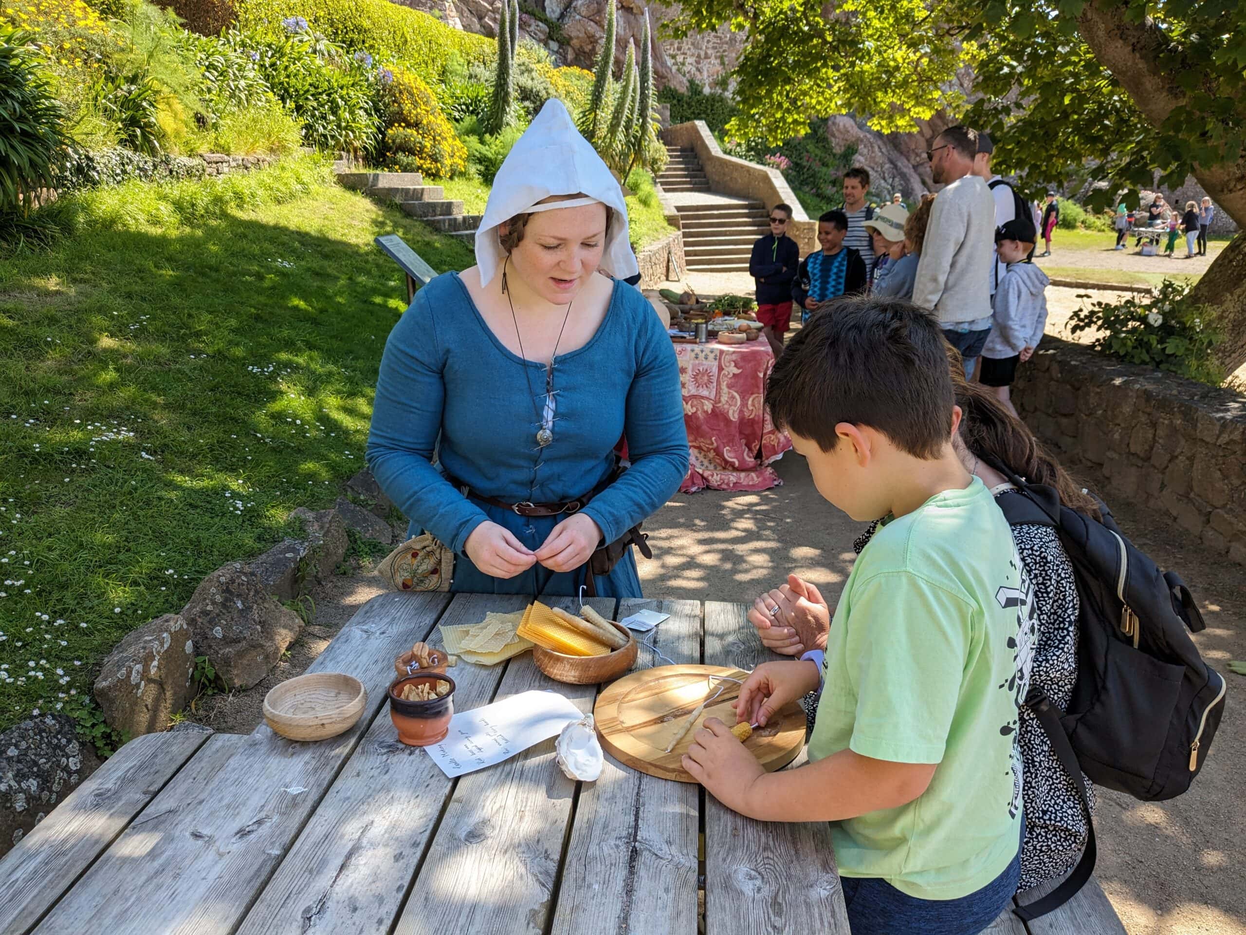 Two children in modern clothes talking to a woman in medieval clothes and holding some beeswax candles.