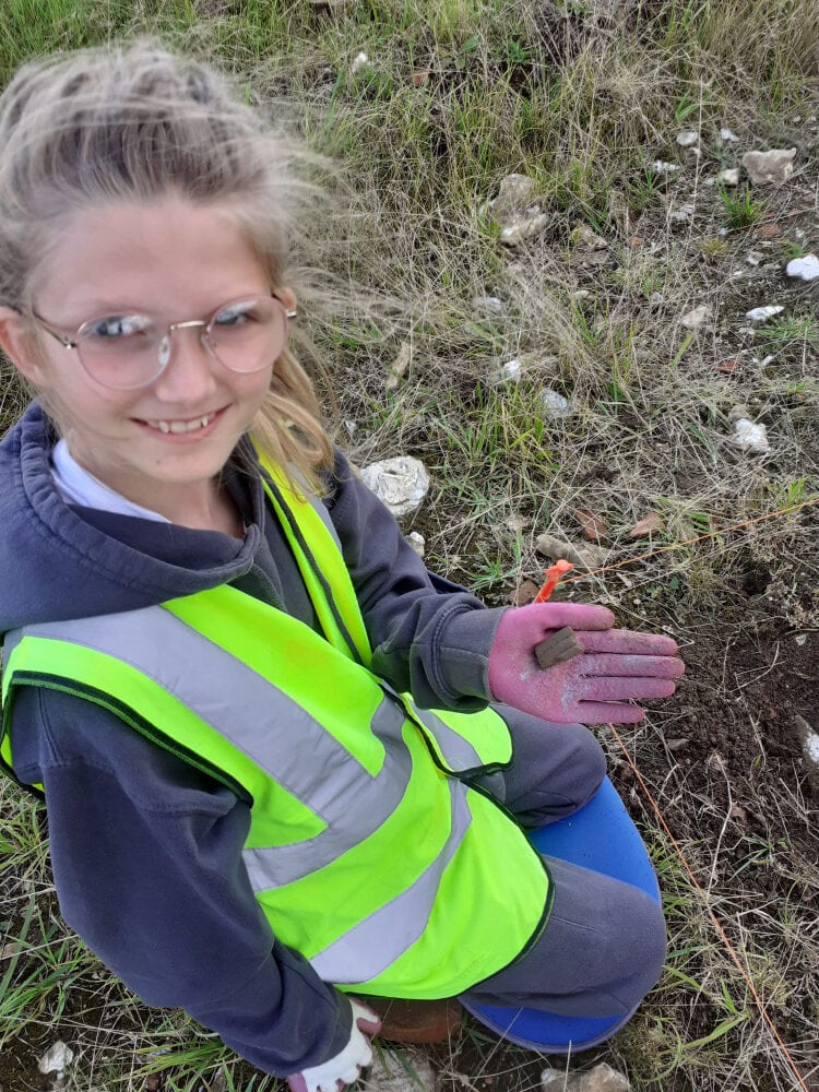 A girl wearing a high-vis vest kneels on the grass holding up a small piece of brown pottery.