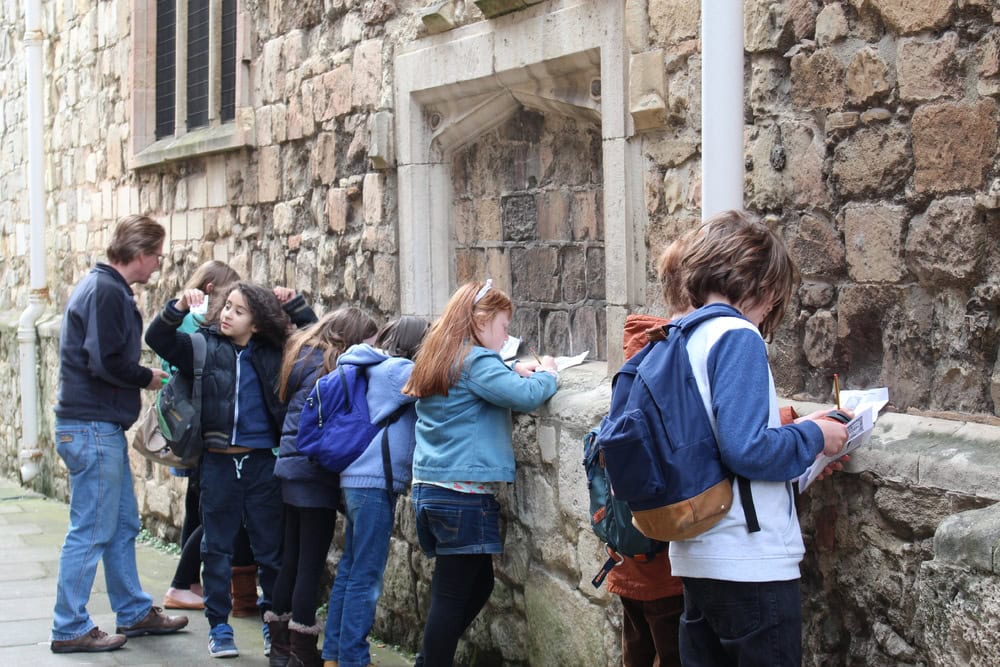 A group of young people leaning against the stone wall of a historic building to write on clipboards.