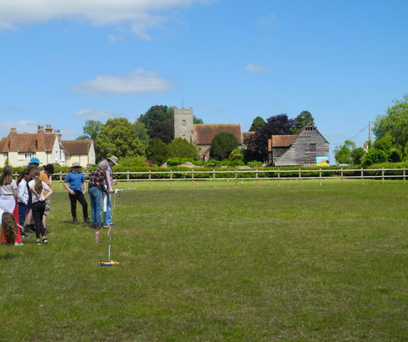 A group of children and adults stand in a field outside a village. One adult is using as Magnetometry machine to take readings.