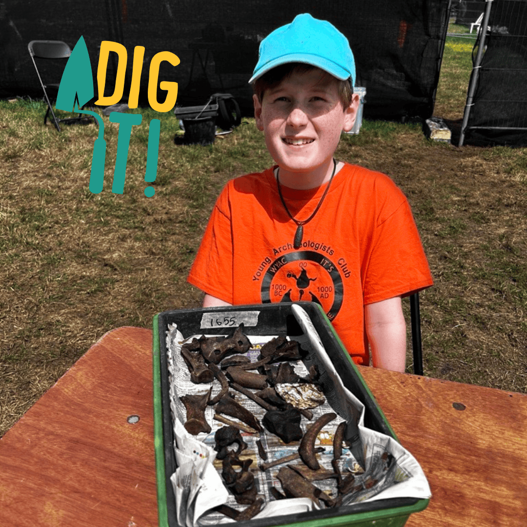 A young person seated at a wooden table on the grass with a tray of excavated animal bones and shells.