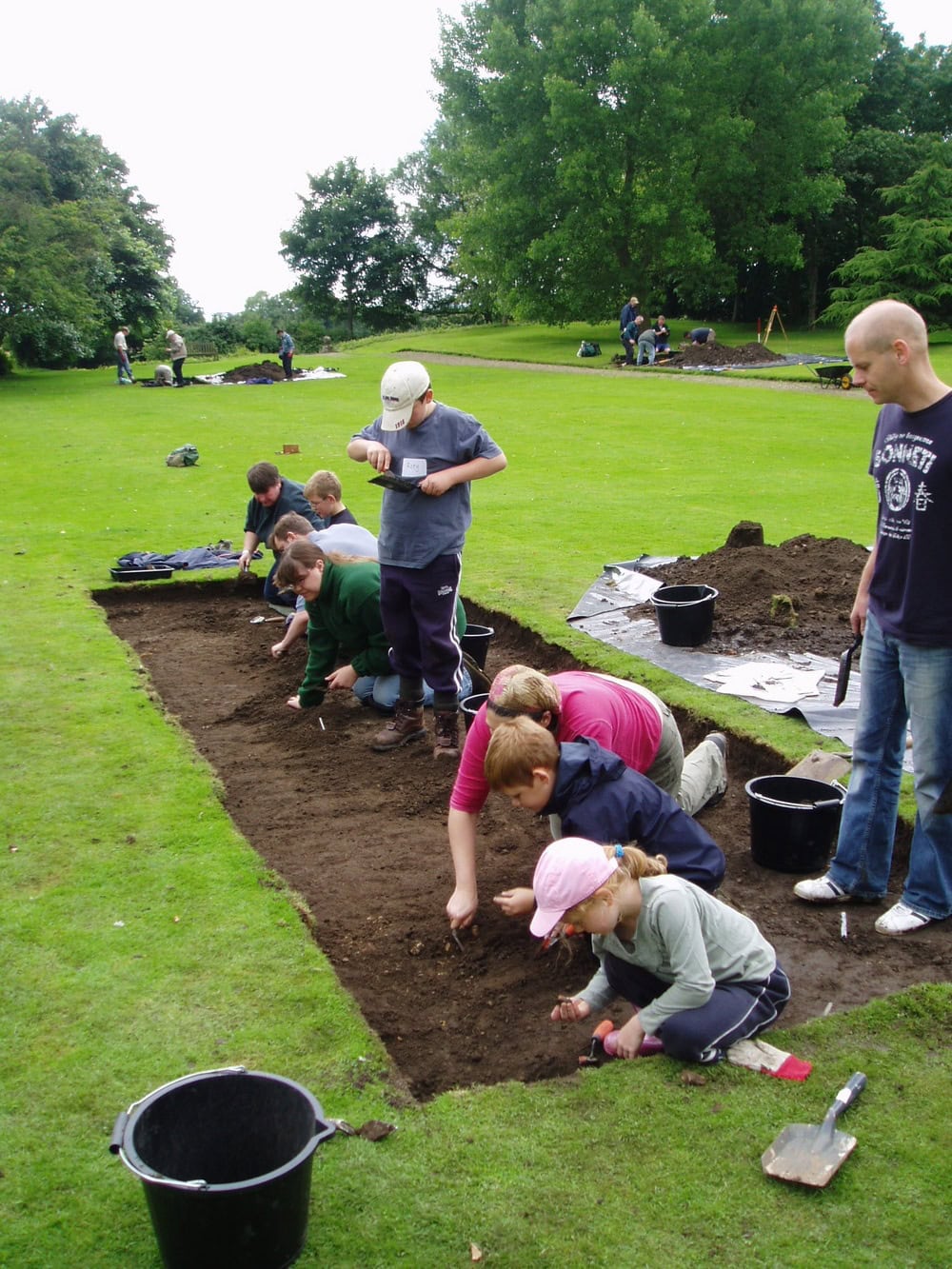 Six young people digging in a long shallow rectangular trench in an archaeological dig.