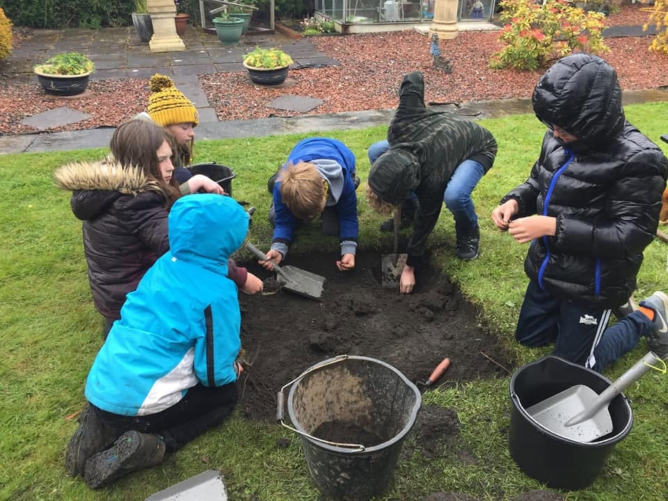 Six young people excavating a shallow square trench on an archaeological dig using metal trowels and buckets.