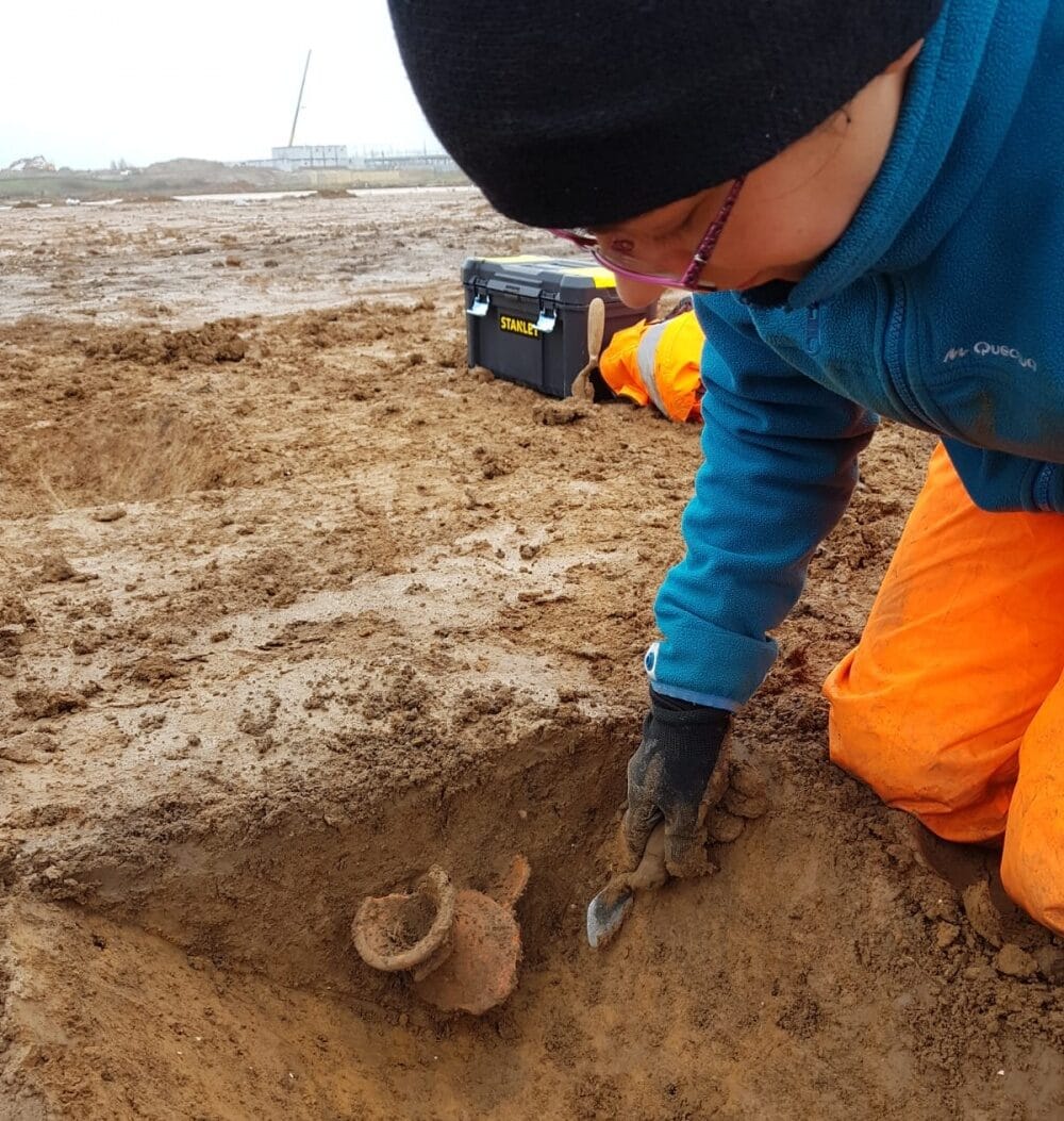 A young person working on an archaeological dig, carefully scraping the soil with a metal trowel to reveal an old pot.