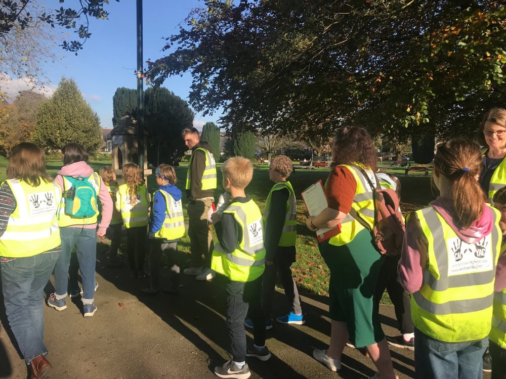 A group of children and adults all wearing high-vis vests with the YAC handprint logo walk through a park on a sunny date.