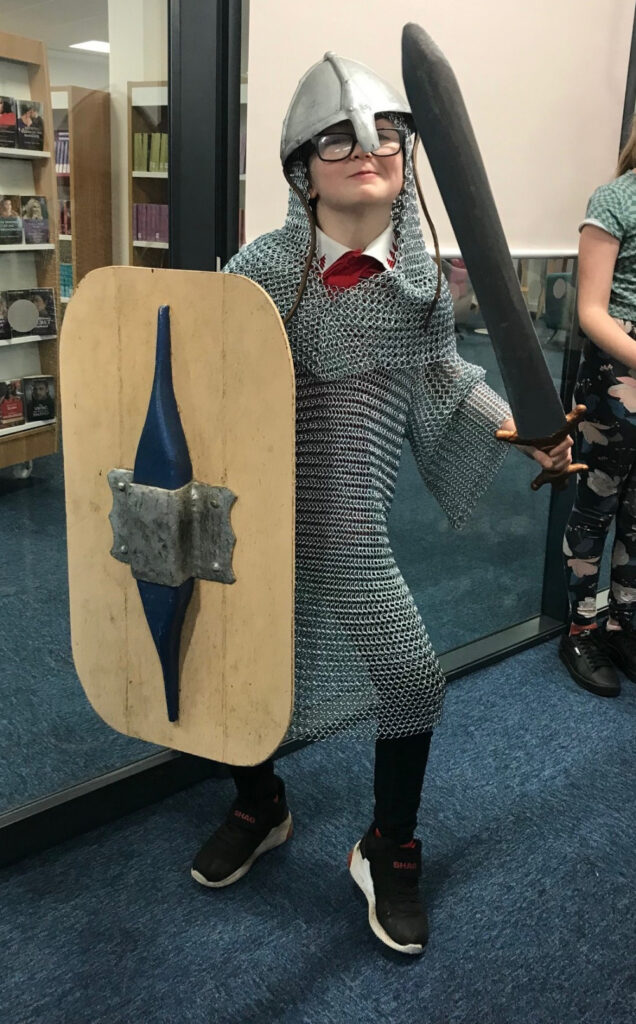 A boy wearing a Norman-style chainmail shirt and a helmet holding a sword and wooden shield.