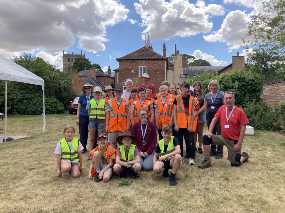 A group of 18 children and 6 adults standing in a field. A gazebo and some buildings are in the background.