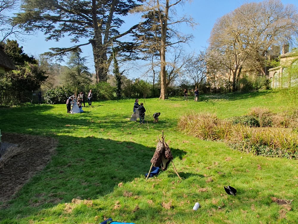 A group of young people in the garden of a museum constructing prehistoric shelters out of sticks and furs.