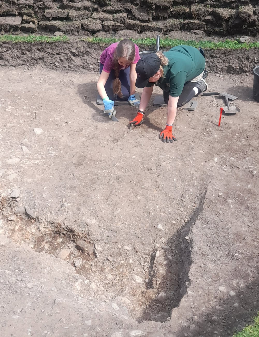 Two girls crouch together digging in an excavation pit.