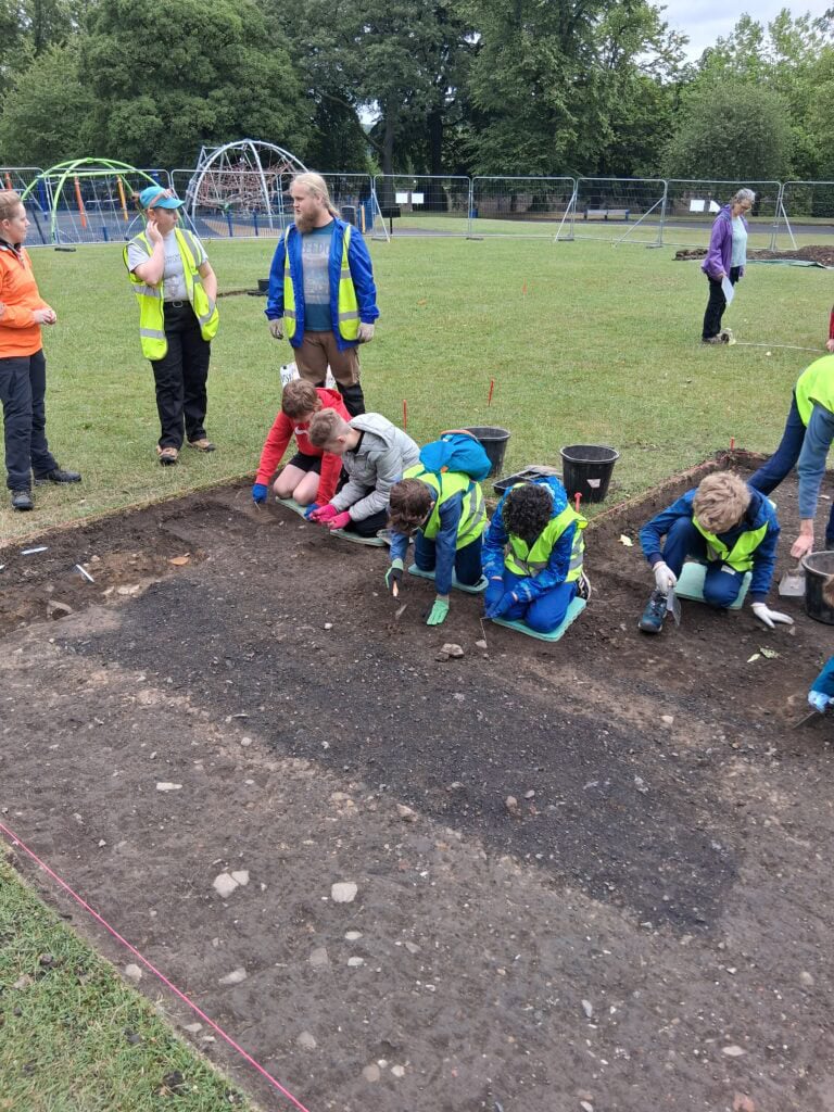 Five children wearing high-vis vests and gloves kneel around the edge of a shallow rectangular archaeological trench, which they excavate with trowels.