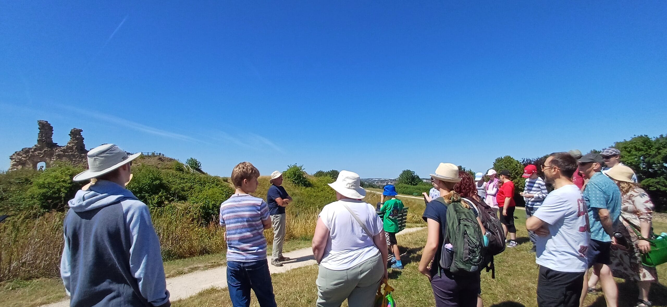 A group of around 20 young people and adults stood on a grassy lawn on a sunny day with castle ruins in the background.