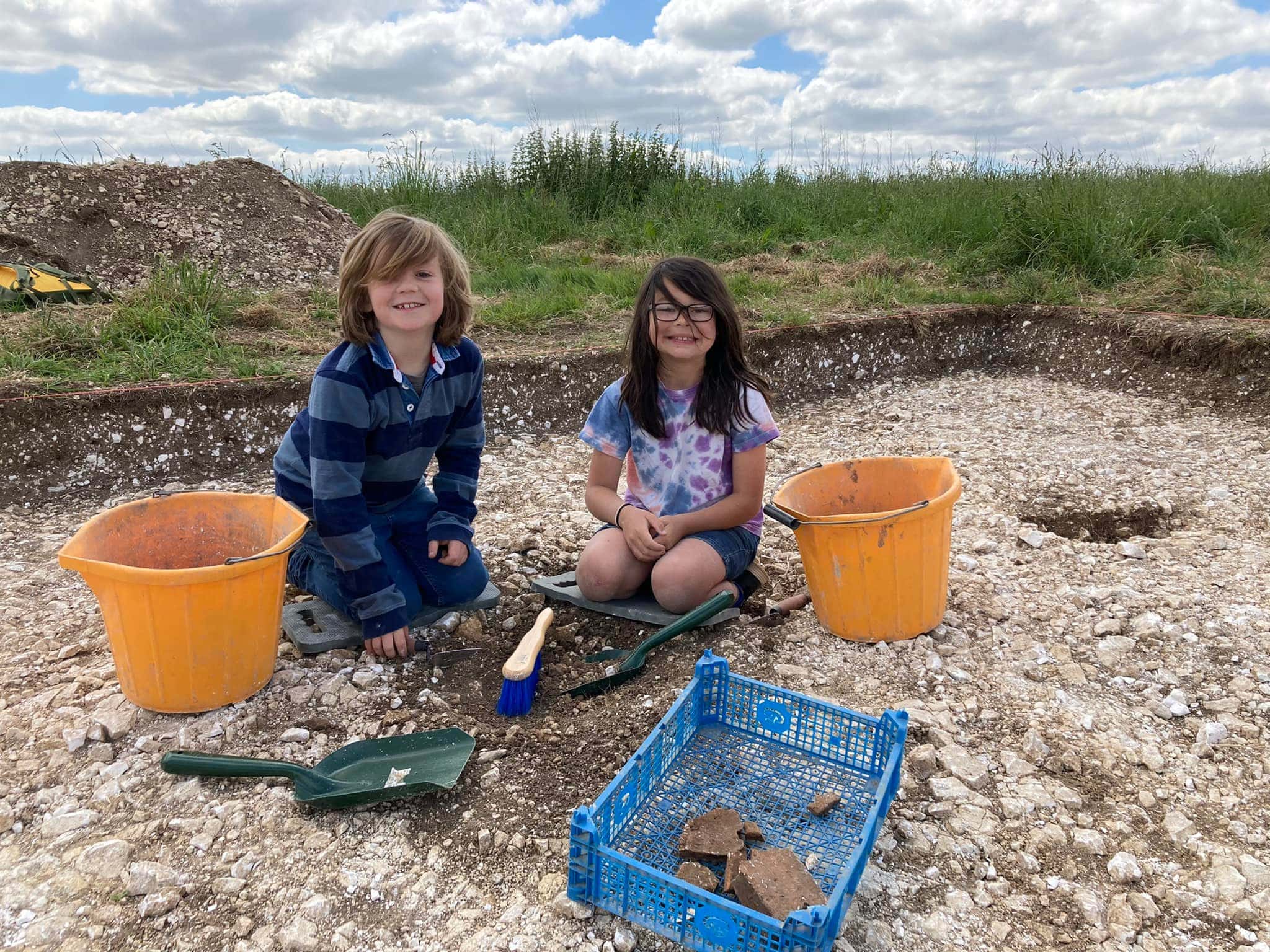 Two young people sat in a trench of an archaeological dig with trowels, brushes, buckets and a tray of finds.