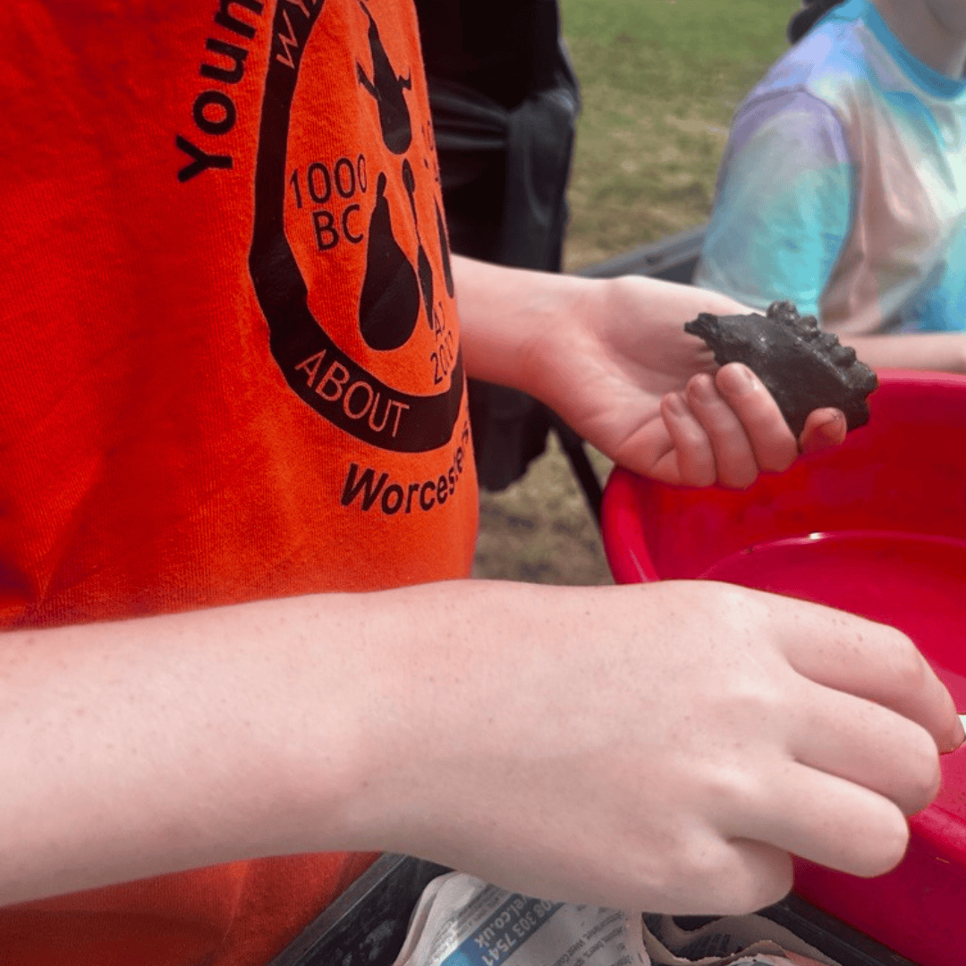 A young person wearing an orange t-shirt washes a piece of muddy pig jaw bone in a red bucket of water.