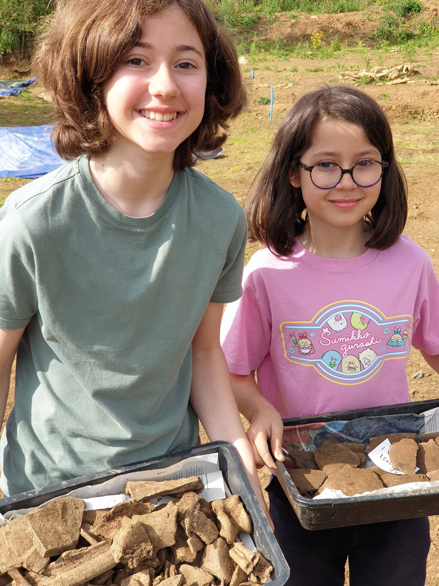 Two young people outside on an archaeological dig site holding trays of finds such as bones and bricks.