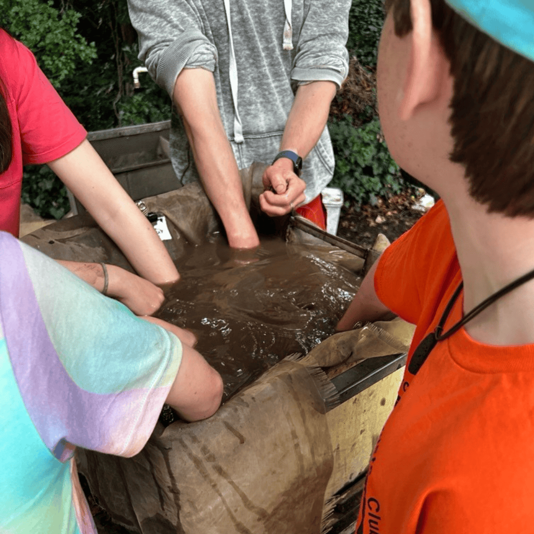 Four young people stick their arms into a big rectangular tray of muddy water.