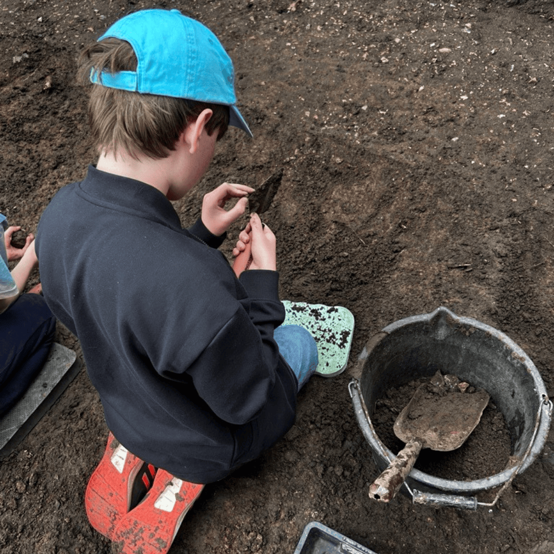 A young person kneels in an archaeological trench and digs with a metal trowel.