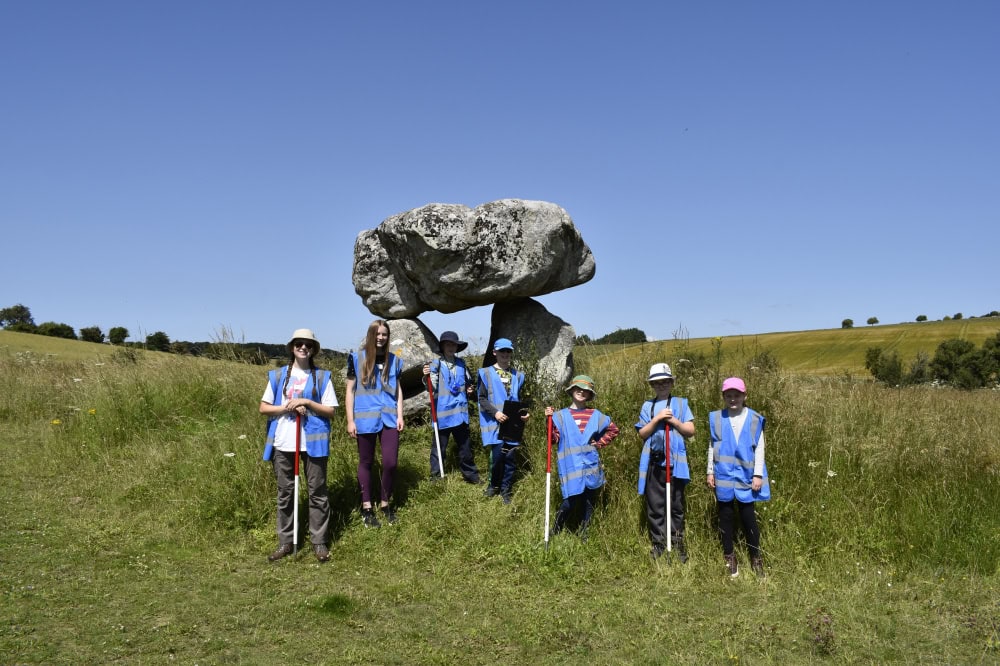 Seven young people wearing blue high-vis vests and holding red and white poles standing in a field on a sunny day in front of a prehistoric arch structure comprising of three huge stones.