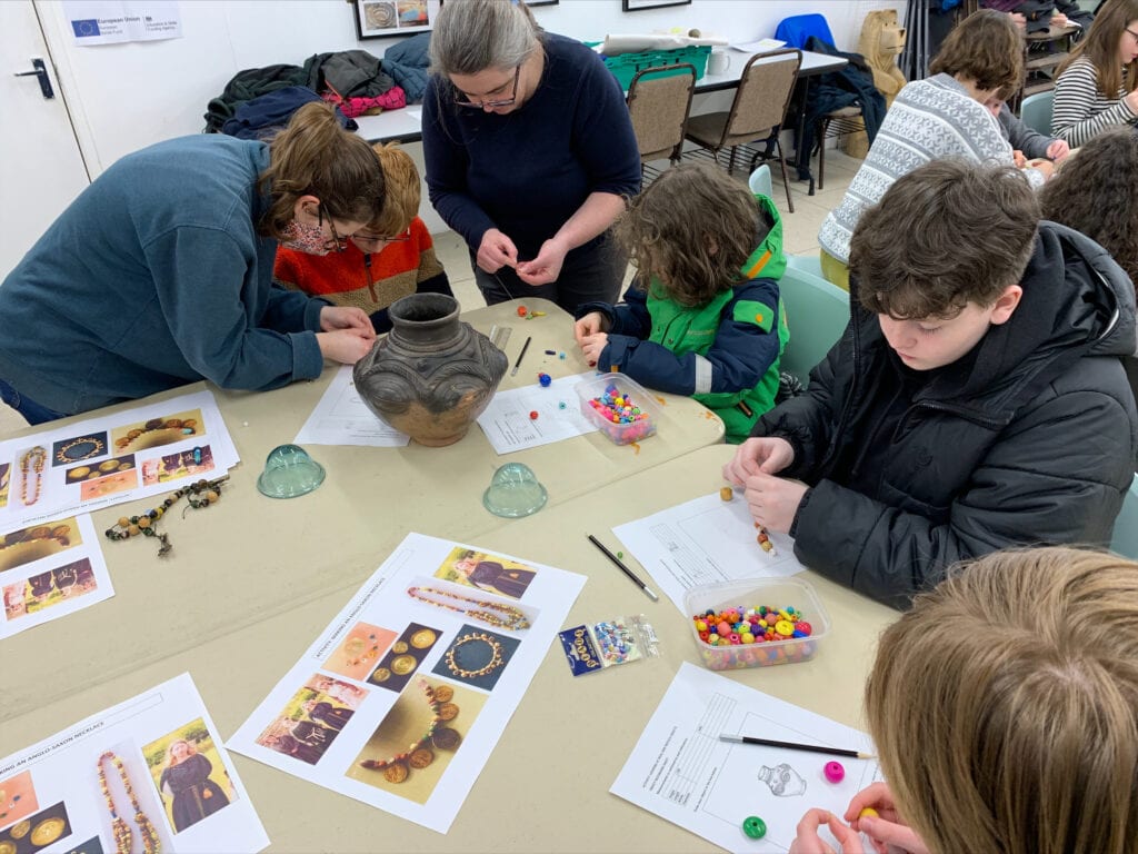 A group of young people sat at a table with bowls of colourful beads and printed photographs of Anglo-Saxon jewellery.
