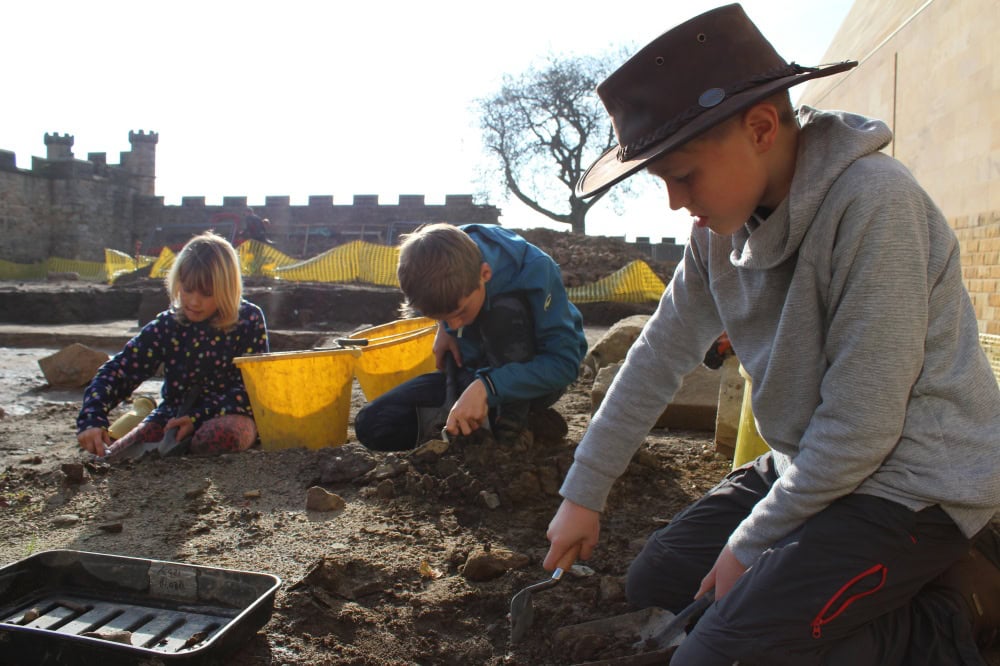 Three young people digging in an archaeological excavation, castle walls are in the background.