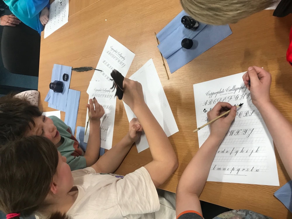 Three children sat at a table doing calligraphy with feather pens and ink.