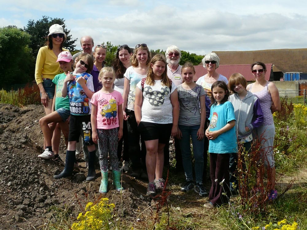 A group of 11 young people and 5 adults standing on an archaeological dig site.