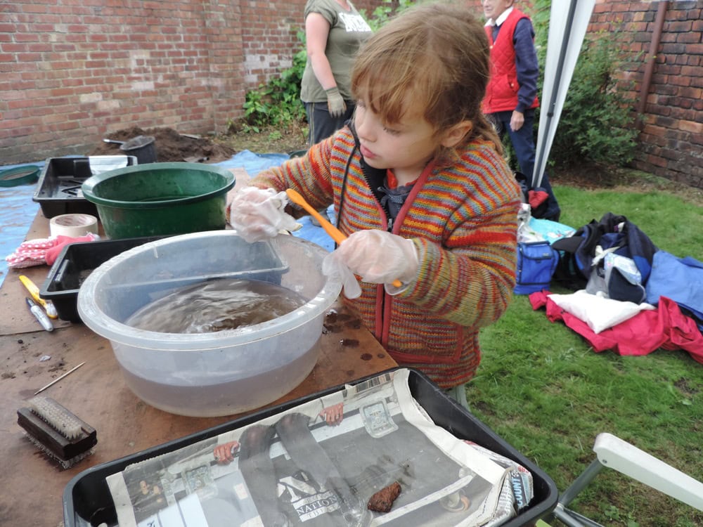 A young person stood at a table outside washing an archaeological find in a washing-up bowl with a toothbrush.