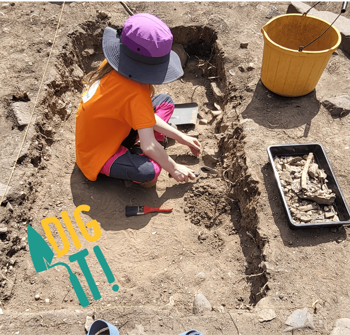 Young archaeologist Abbie using a trowel to excavate in a shallow trench next to a tray of animal bones and a yellow bucket of spoil. The text reads 'Dig It!' with an illustration of a trowel.