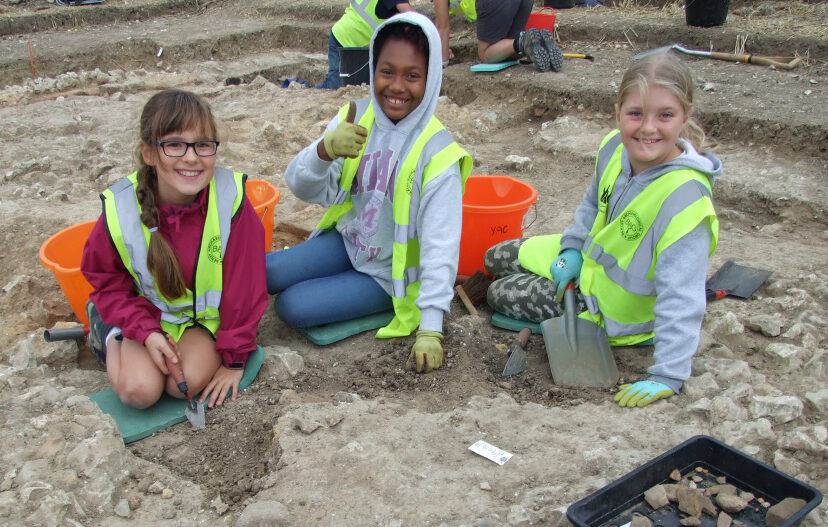 Three young people wearing high-vis vests digging on an archaeological excavation.