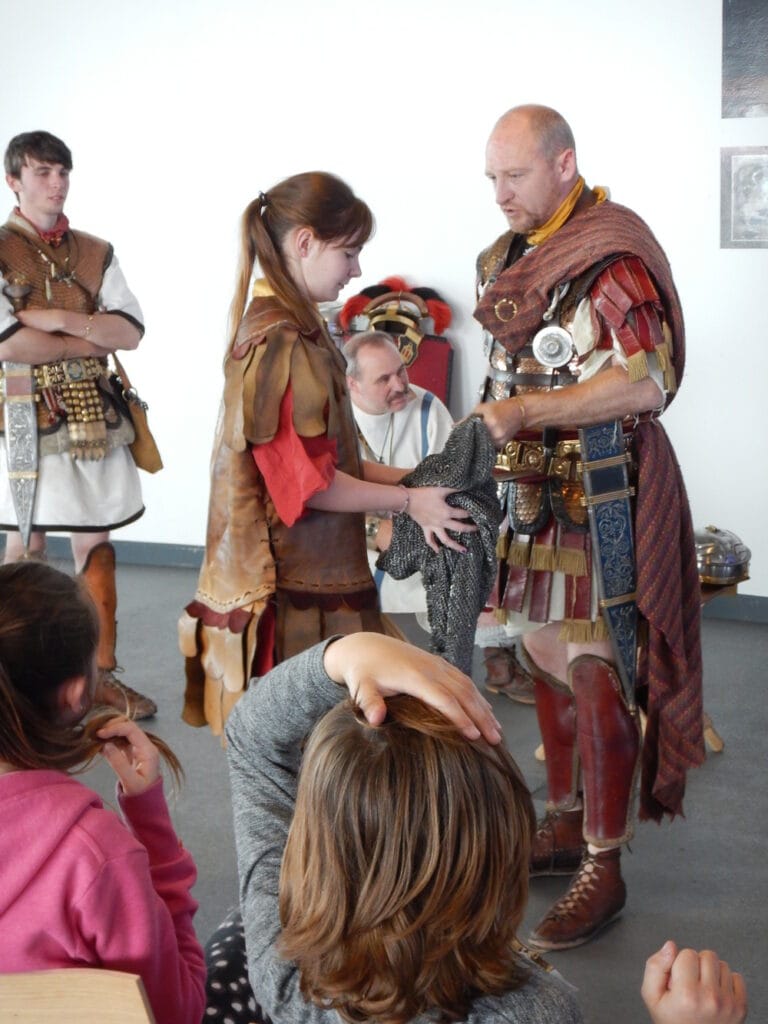 Two adults dressed as ancient Roman soldiers helping a young person wearing a Roman tunic hold a chain-mail shirt. Other young people watch.