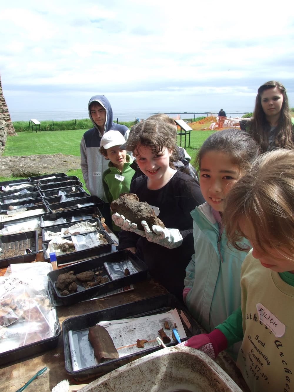 Six young people stand outside in front of a table with trays of archaeological objects such as animal bones and building materials.