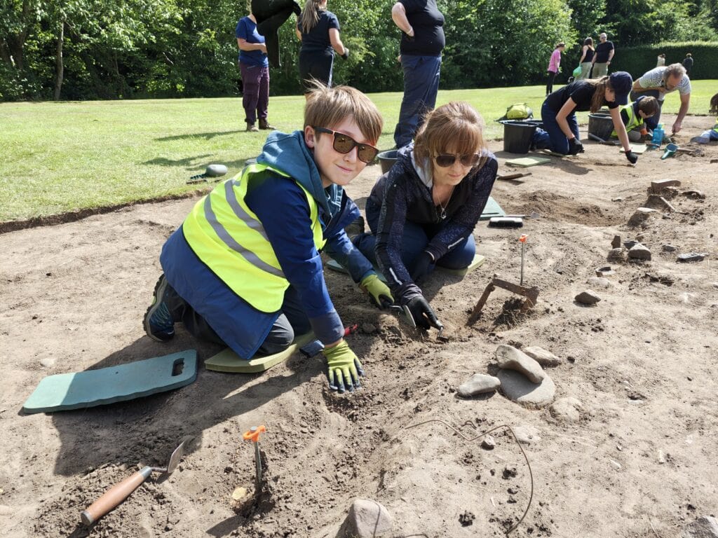 A young person wearing a high-vis vest and gloves kneels in an archaeological trench and scrapes the soil with a trowel. Other adults and young people excavate the site in the background.