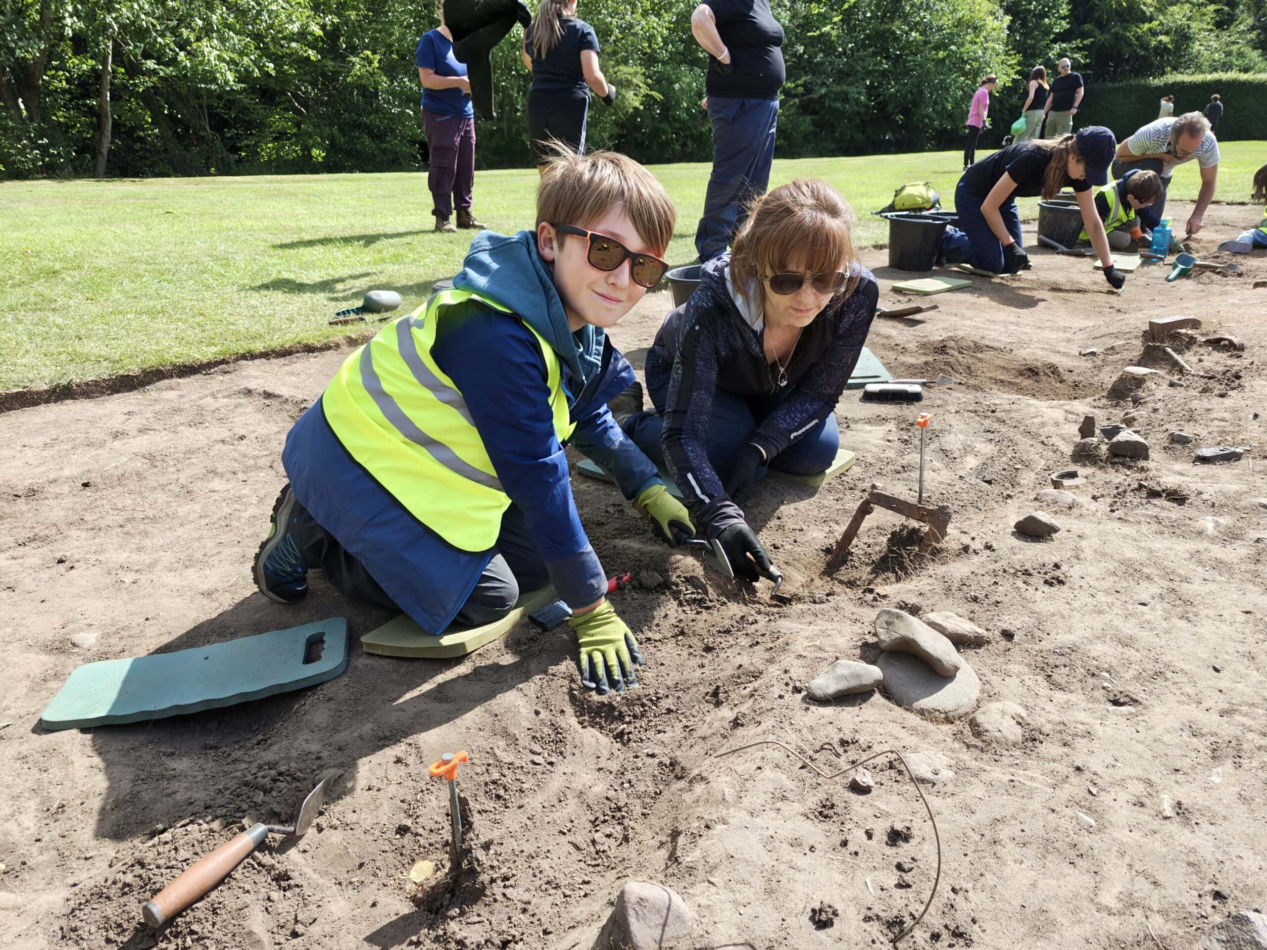 A young person wearing a high-vis vest and gloves kneels in an archaeological trench and scrapes the soil with a trowel. Other adults and young people excavate the site in the background.