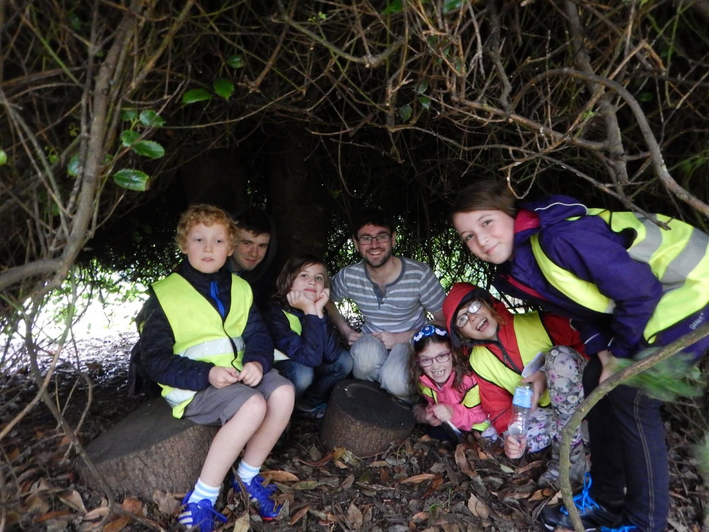 Six young people and one adult crouch in an enclosure of trees.